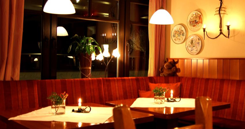 Wirtschaft des Restaurants. Draußen ist es schon dunkel, innen sind Kerzen an. Das Mobiliar ist aus hellem Holz mit roten Bezügen.