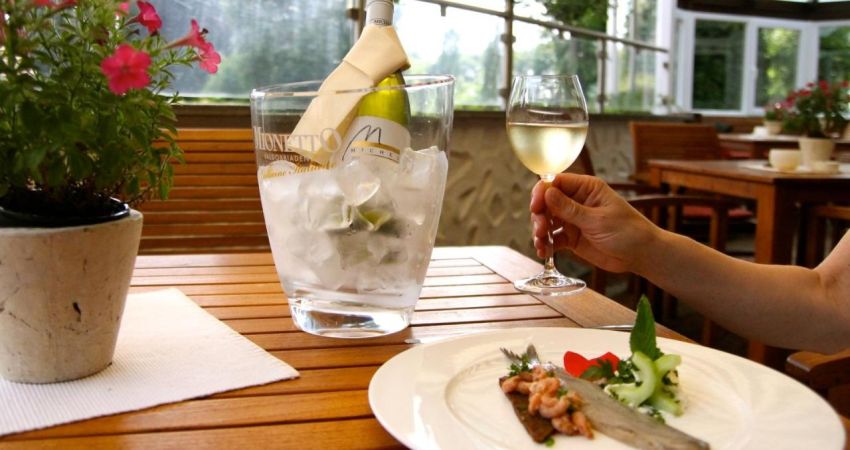 Ein Tisch aus Holz auf der Terrasse. Gedeckt mit einer Flasche Weißwein und einem Tellergericht auf dem Matjes , Krabben und Pumpernickel angerichtet sind. 