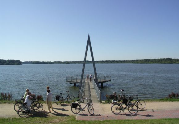 Aussichtsplattform an der Thülsfelder Talsperre: Brücke mit A-förmigem Bauwerk, Fahrräder und Personen am Ufer.